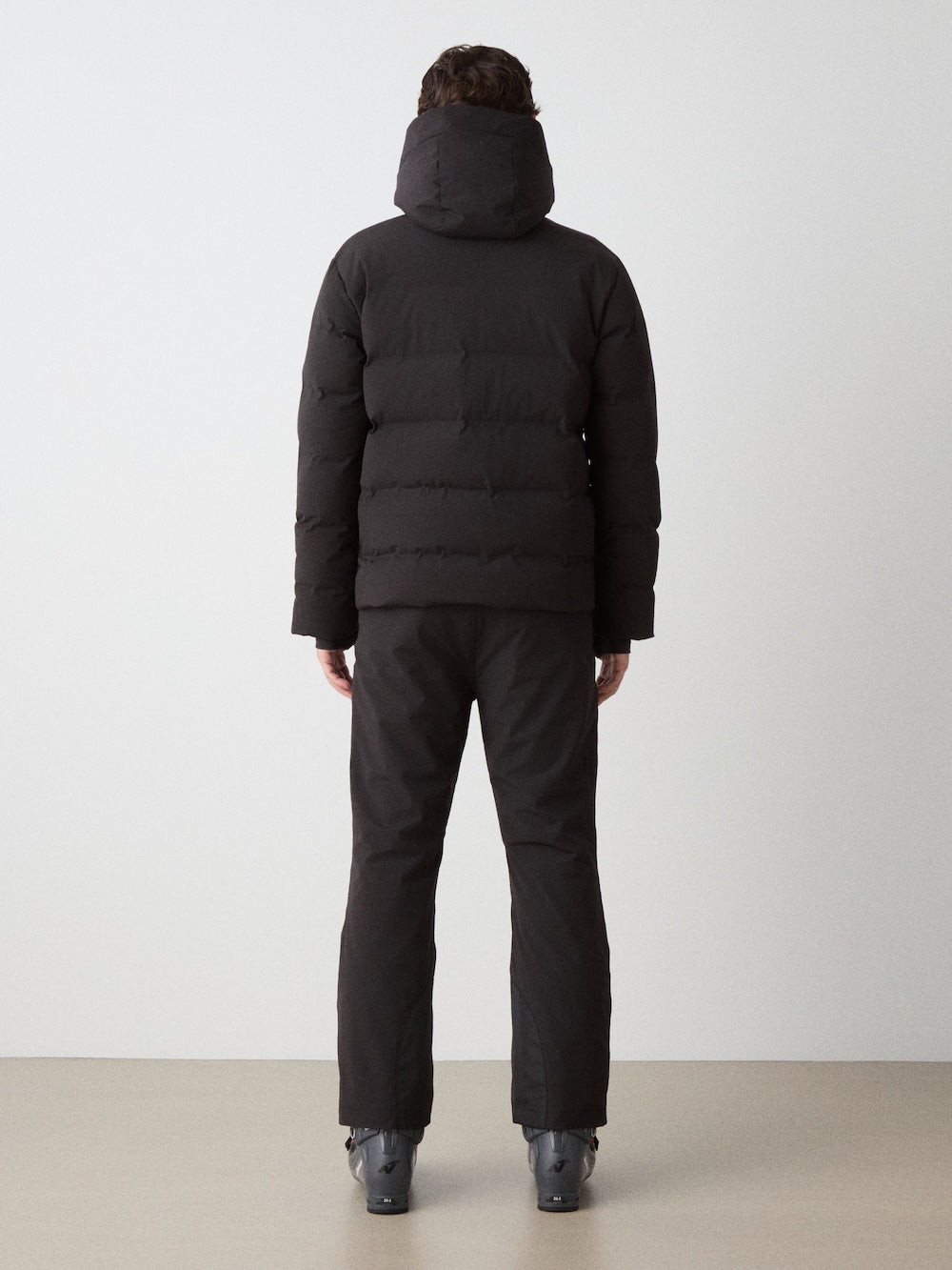 A person stands indoors facing away, wearing the We Norwegians Nordlys Ski Jacket for men—black, waterproof, hooded—paired with black pants and boots on a neutral floor against a plain white background.