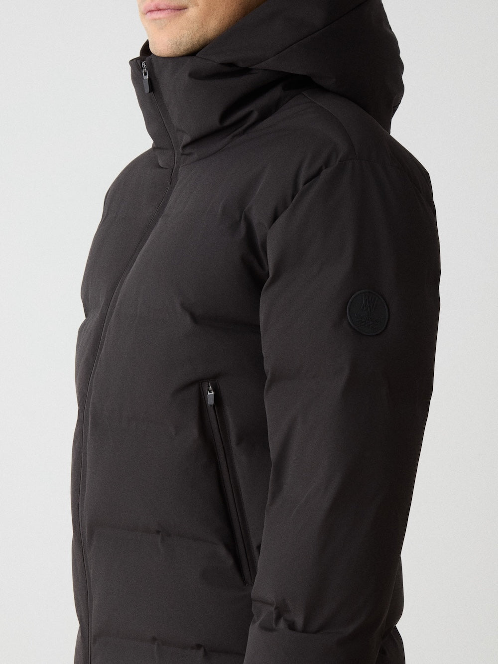 A person wearing the We Norwegians Nordlys Ski Jacket for men in black, zipped up with hood on, is shown from the side with their face partially out of frame; the jacket features zippered pockets and a circular patch on the upper sleeve.