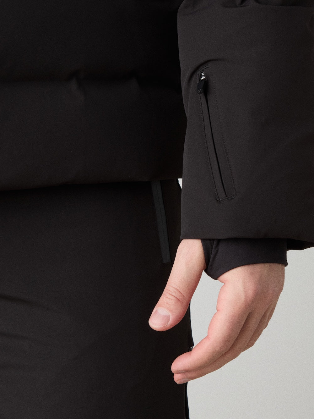 A close-up of a person in the We Norwegians Nordlys Ski Jacket men, featuring responsible down, waterproof finish, a zippered sleeve pocket, and cuff thumbhole, with part of their hand and black pants visible.