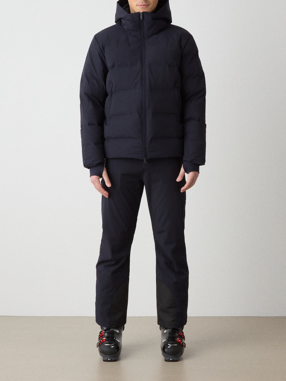 A person stands facing forward in a We Norwegians Nordlys Ski Jacket for men—black, hooded, waterproof, and filled with responsible down—paired with black pants and black boots with red accents against a plain light-colored background.