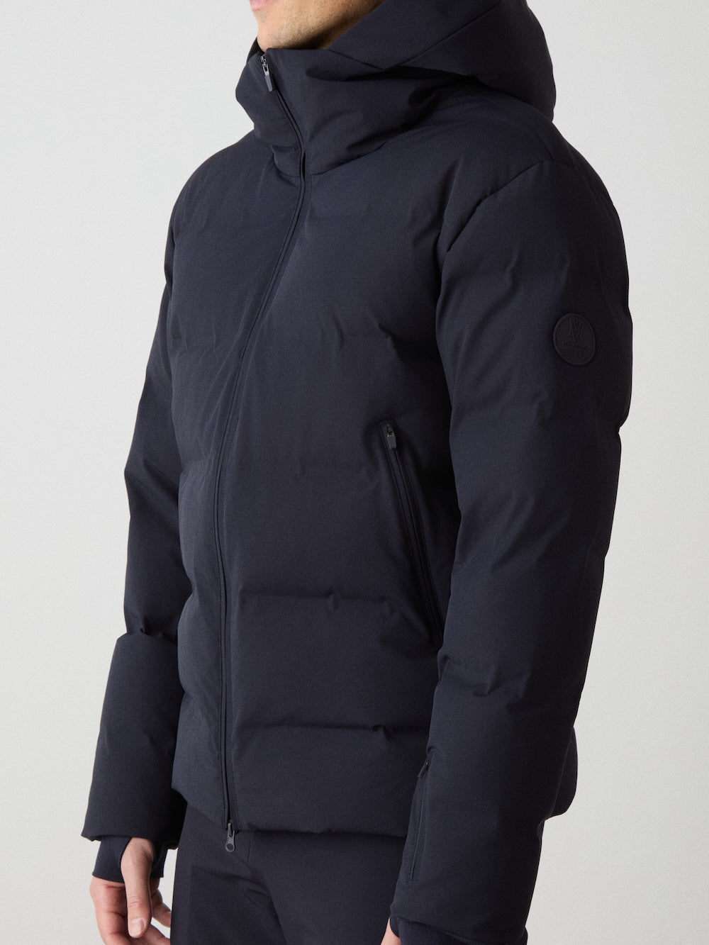 A man stands against a plain light background wearing the We Norwegians Nordlys Ski Jacket, a dark, hooded waterproof puffer with zipper, side pockets, responsible down fill, and a round patch on the upper sleeve. His face is partially out of view.