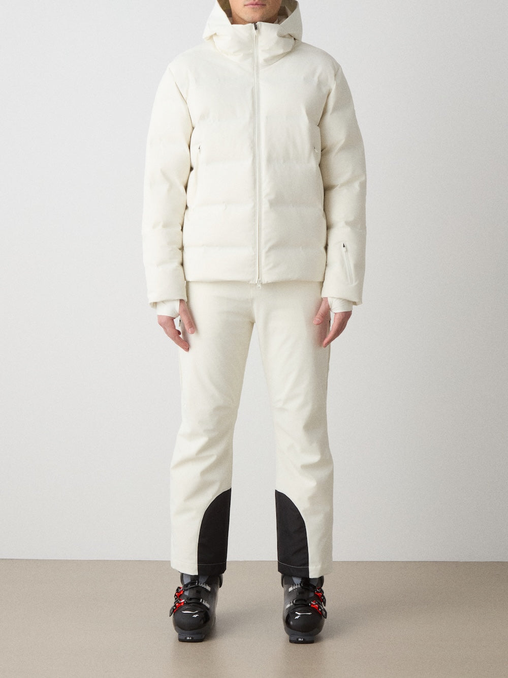 A person in an all-white winter outfit, featuring the We Norwegians Nordlys Ski Jacket for men and fitted pants with black ankle patches, paired with black ski boots, stands against a plain background.