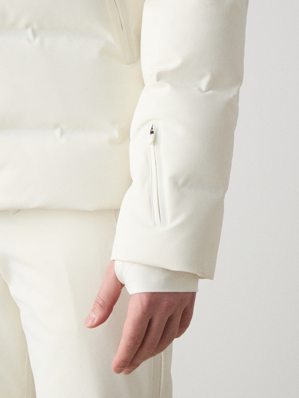 A person stands against a plain light background, wearing the We Norwegians Nordlys Ski Jacket men in white; the focus is on the sleeve’s small zippered pocket and their relaxed hand.