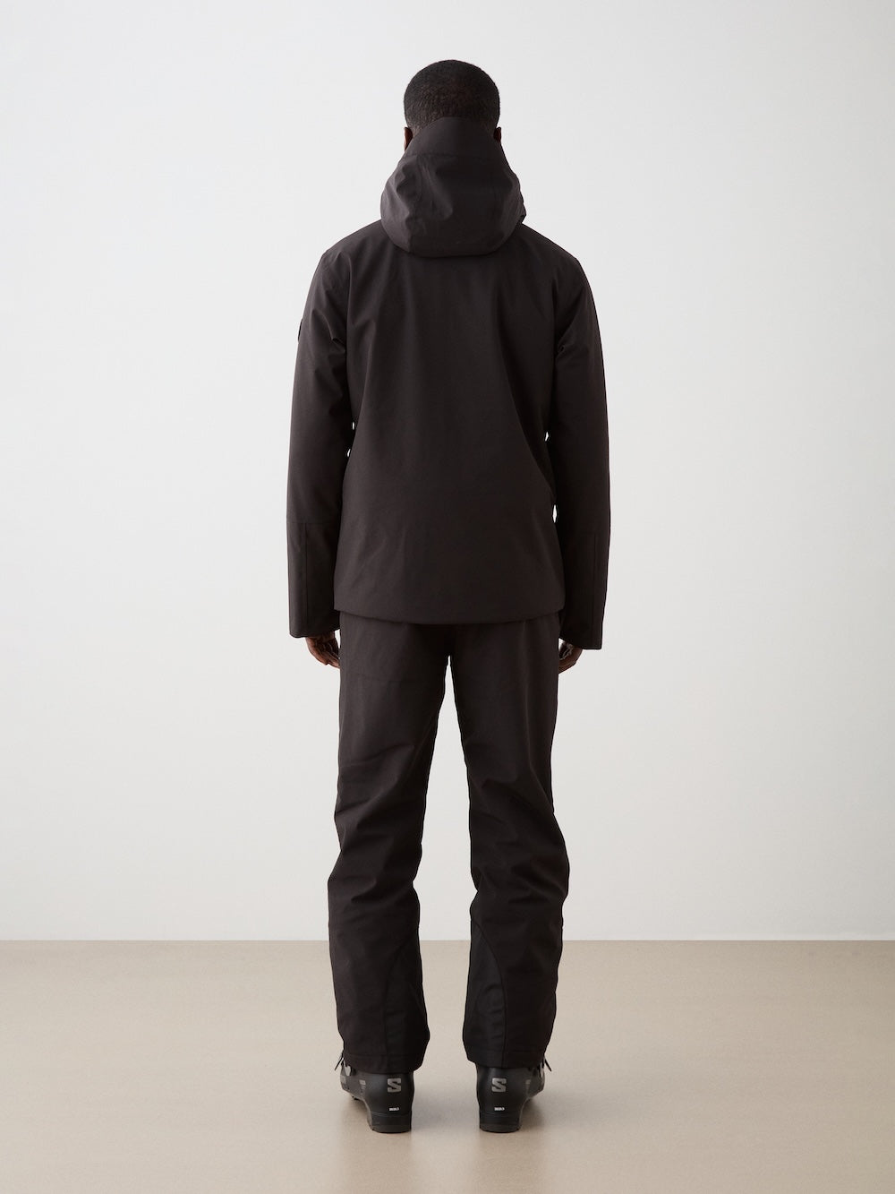 A man faces away from the camera, wearing a black We Norwegians Hammerfest Ski Jacket for men with matching pants and boots, set against a plain light background.