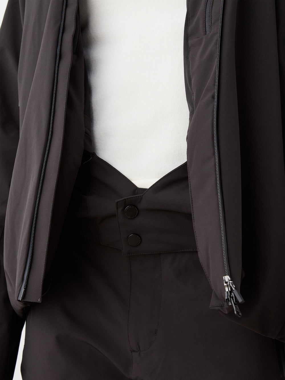 A close-up shows a person wearing the We Norwegians Hammerfest Ski Jacket for men, unzipped to reveal a white shirt and black pants with a unique angular waistband closed by two black buttons.