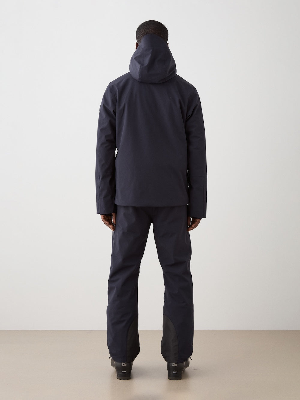A person faces away from the camera wearing a dark We Norwegians Hammerfest Ski Jacket for men with matching pants and boots, set against a plain light background.