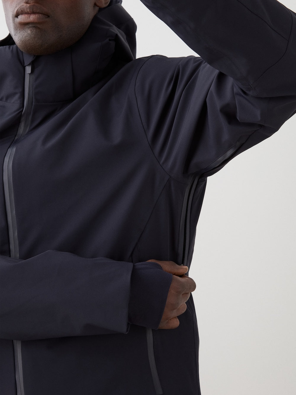A person wears a dark, hooded We Norwegians Hammerfest Ski Jacket men, raising their left arm and adjusting the sleeve. Featuring thumbholes and a sleek, minimalist look, the jacket stands out against a plain, light-colored backdrop.