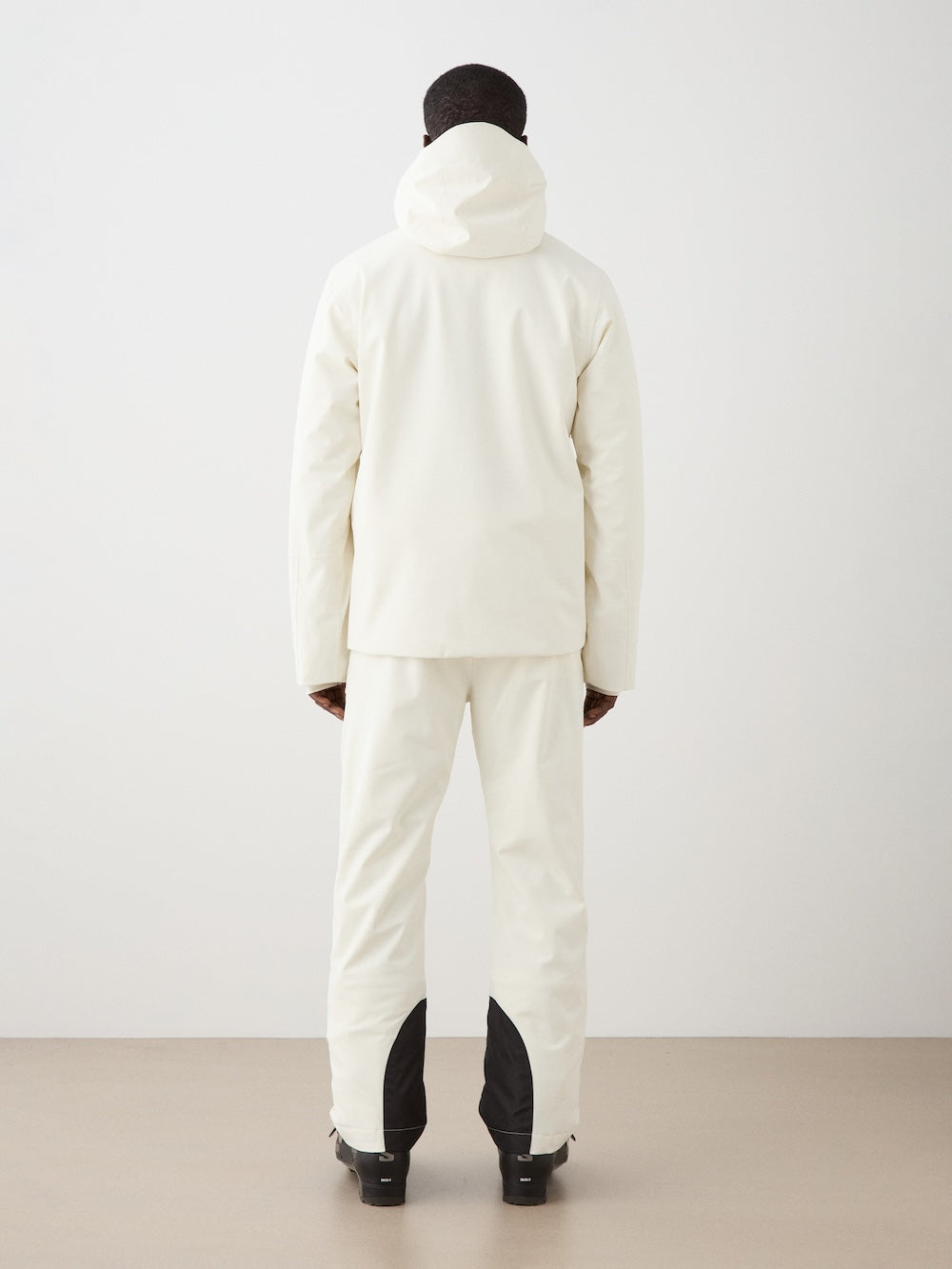 A man faces away from the camera, wearing a white We Norwegians Hammerfest Ski Jacket and matching white pants with black lower leg patches, set against a plain light background.