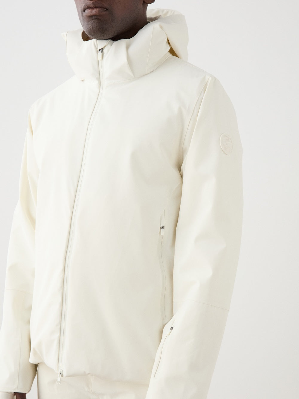 A man wears the We Norwegians Hammerfest Ski Jacket in off-white, posing against a white background. The hooded zip-up features a subtle sleeve logo and a minimalist, 4-way stretch design.