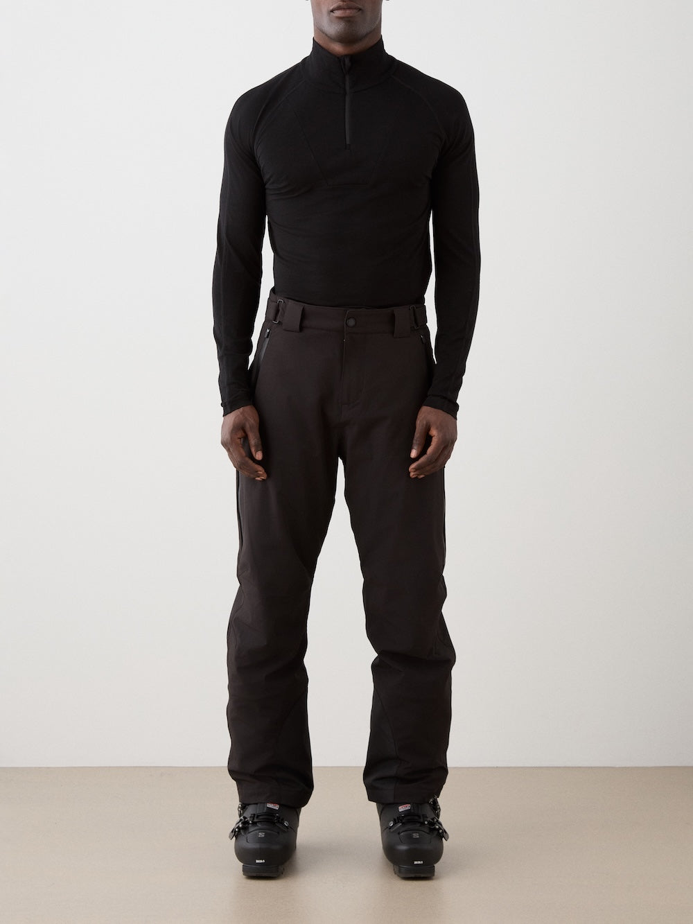 A person stands facing forward, wearing a fitted black long-sleeve top, We Norwegians Hammerfest Ski Pant men, and black ski boots against a plain, light-colored background.