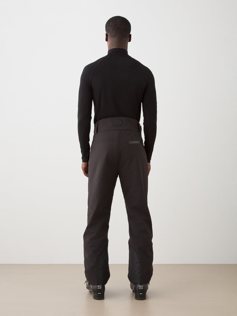 A person stands facing away from the camera wearing a fitted black long-sleeve top, black boots, and We Norwegians Hammerfest Ski Pant men, against a plain light background.