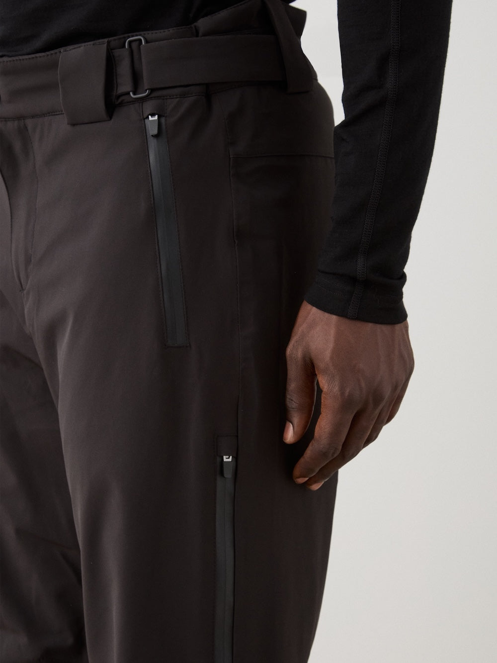 A person is shown wearing We Norwegians Hammerfest Ski Pant men—dark, waterproof pants with sleek zipper details—paired with a long-sleeve black shirt, highlighting the modern style and quality fit around the lower torso.