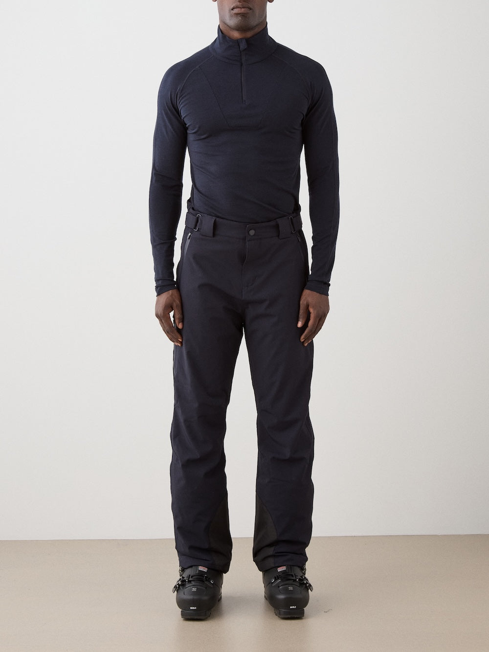 A person stands facing forward, wearing a fitted, long-sleeve, dark navy top and We Norwegians Hammerfest Ski Pant men with a belt and black ski boots, set against a plain, light-colored background.