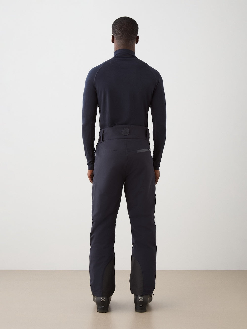 A man faces away from the camera, wearing a fitted black top, We Norwegians Hammerfest Ski Pant men, and black ski boots against a plain light background.