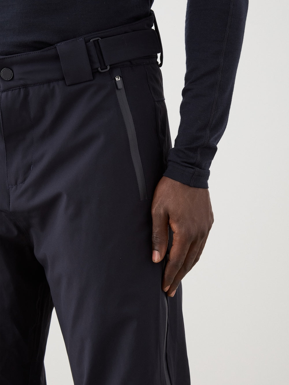 A person wearing We Norwegians Hammerfest Ski Pant men in black, featuring a zipper pocket, and a matching black long-sleeve top stands with their right hand resting along their thigh.