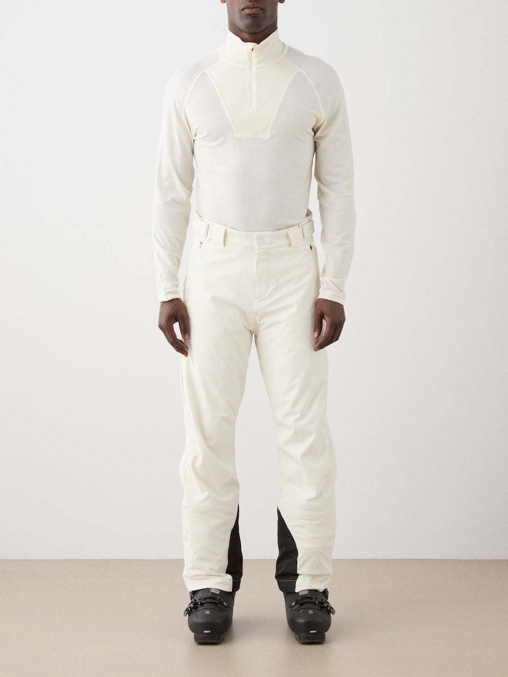 A person stands facing forward, wearing a fitted, light zip-up top and We Norwegians Hammerfest Ski Pant men with black lower leg patches, paired with black ski boots, against a plain background.