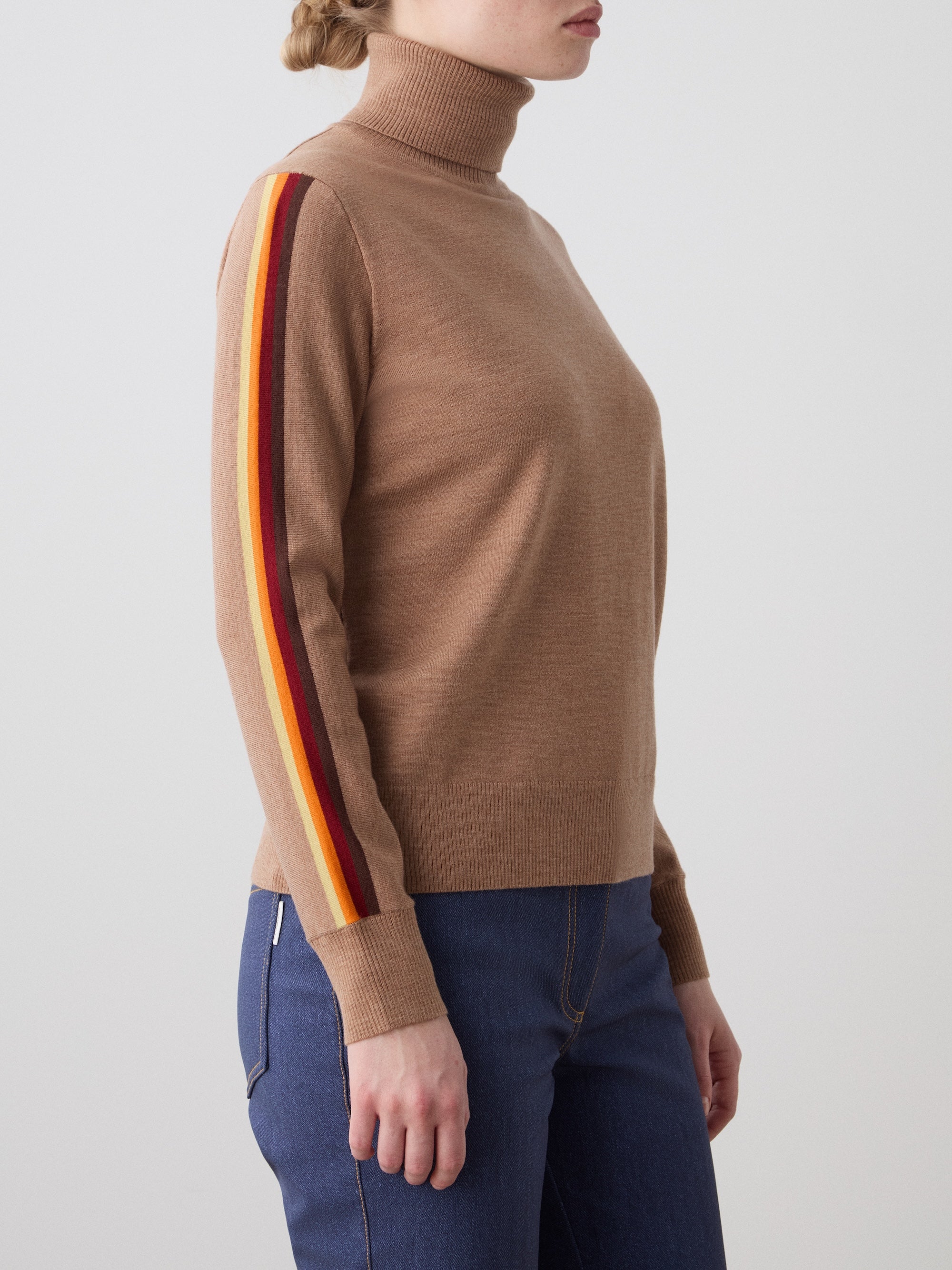 A person wears the We Norwegians FreeBird Turtleneck women—a tan merino wool sweater with red, yellow, and orange sleeve stripes—paired with blue jeans, standing against a plain light background.