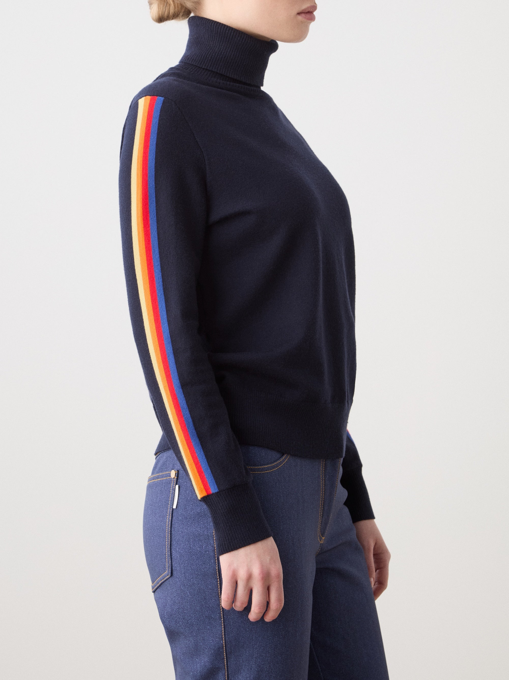 A person models the We Norwegians FreeBird Turtleneck for women—navy merino wool with red, orange, and yellow sleeve stripes—paired with blue jeans, standing sideways against a plain background.