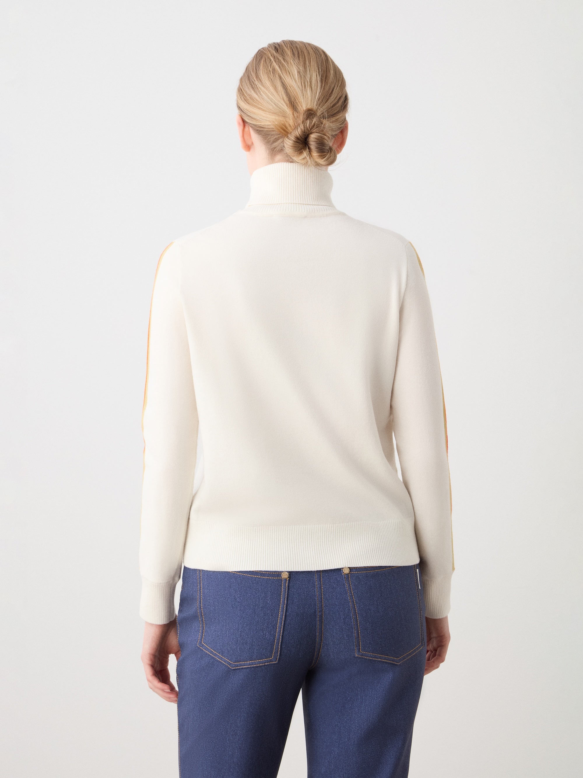 A woman with blonde hair in a low bun is seen from behind wearing the We Norwegians FreeBird Turtleneck women’s sweater—white merino wool with yellow sleeve stripes—and blue jeans with contrast stitching, standing against a plain light background.