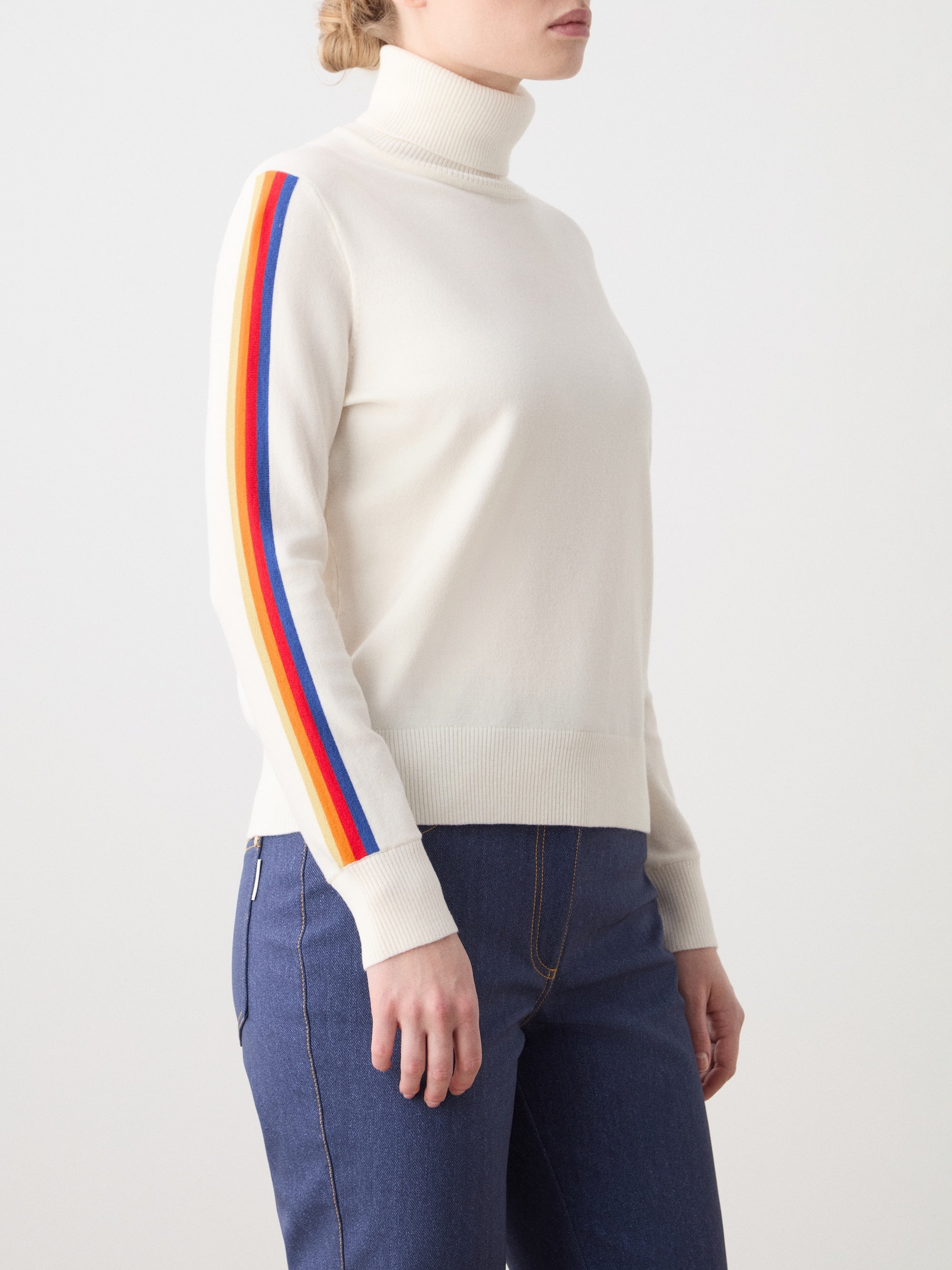 A woman wears the We Norwegians FreeBird Turtleneck, featuring red, yellow, and blue sleeve stripes, paired with blue jeans, standing against a plain light background.