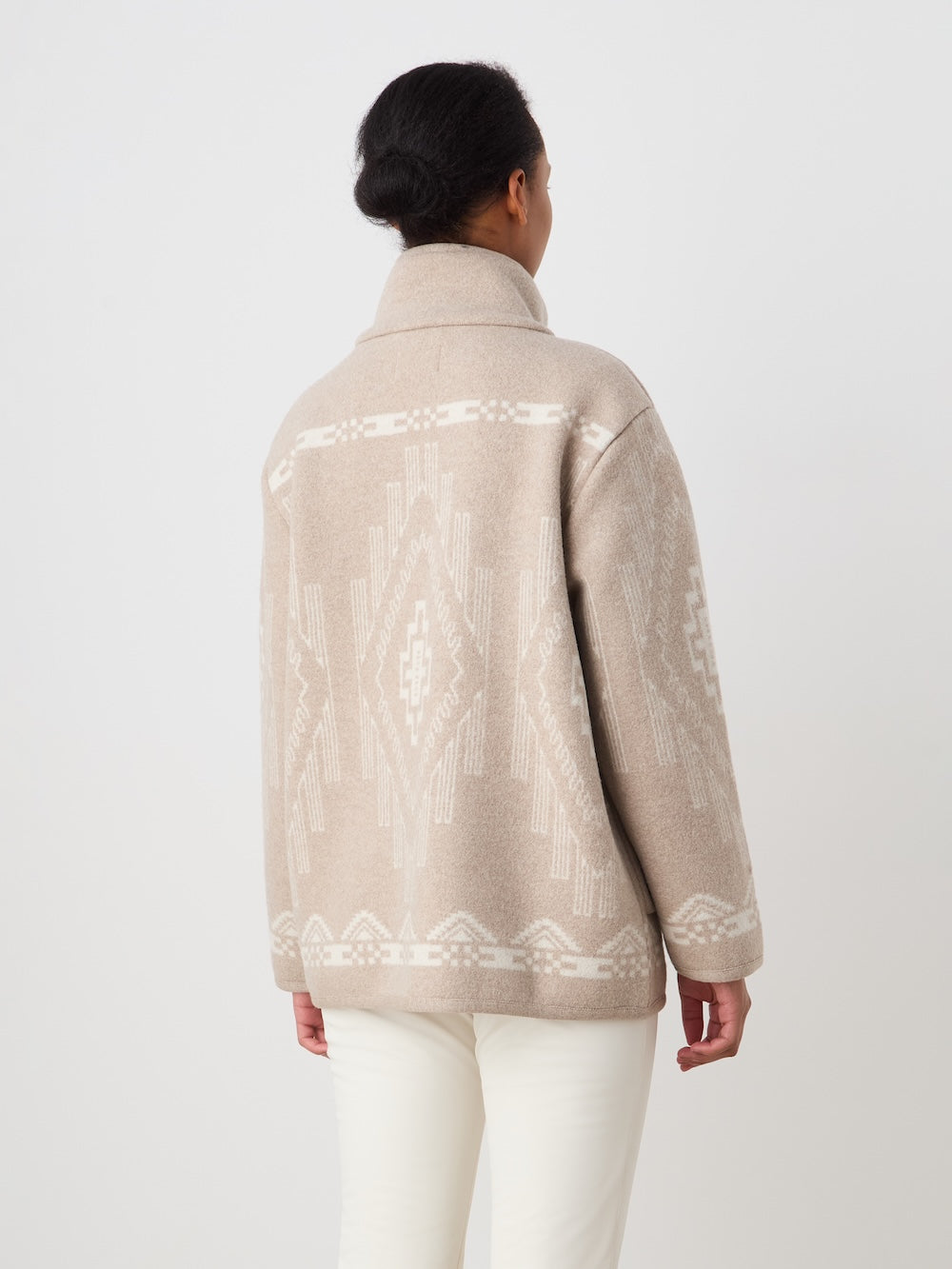 A person with dark hair pulled back is seen from the back wearing the We Norwegians Pioneer Jacket women - Pre Spring 26, featuring a beige color and white geometric pattern, paired with off-white pants against a plain white background.