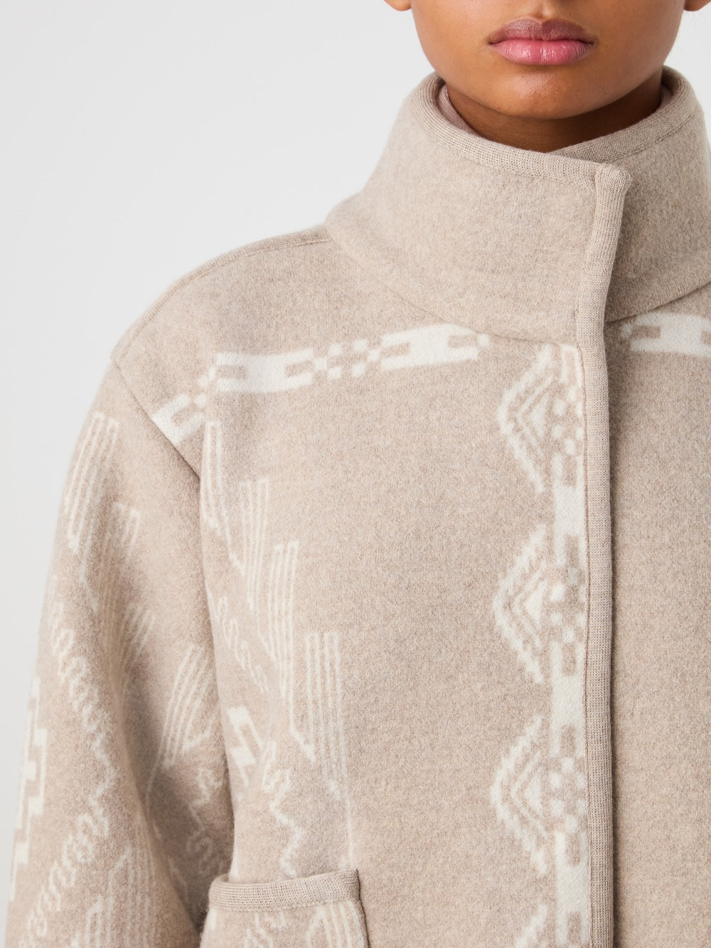 Close-up of a woman wearing the We Norwegians Pioneer Jacket women - Pre Spring 26, a beige wool coat with white geometric patterns, high collar, and front pockets; only her lower face and upper body are visible.