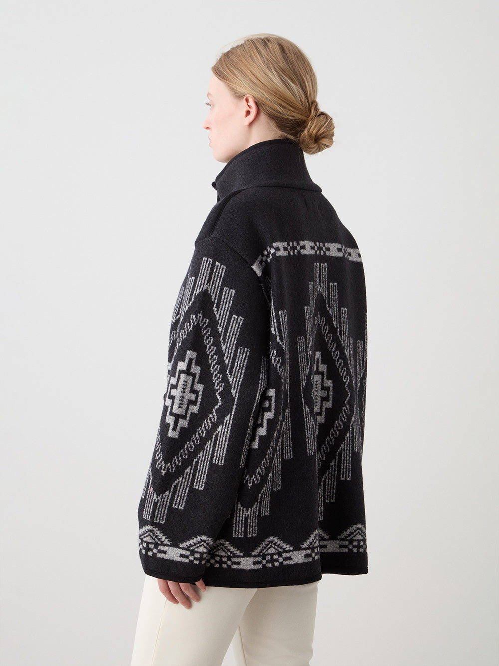 A woman with light skin and blonde hair in a low bun wears the We Norwegians Pioneer Jacket, an oversized black and gray patterned boiled wool piece with geometric designs, standing sideways against a plain, light background.
