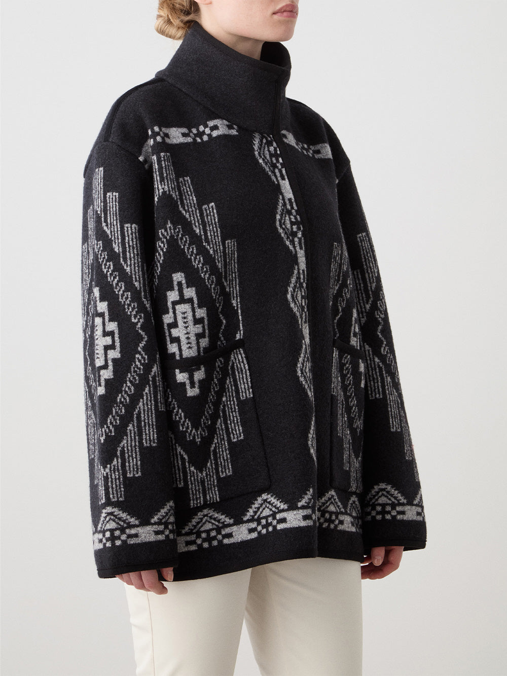 A woman wears the We Norwegians Pioneer Jacket—black and gray boiled wool with geometric patterns, oversized fit, high collar, and long sleeves—paired with white pants against a plain light background.