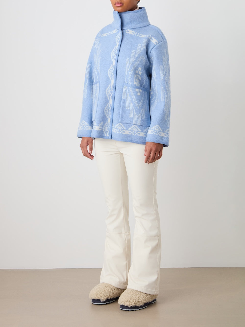 A person stands indoors against a white wall and tan floor, wearing the We Norwegians Pioneer Jacket women - Pre Spring 26 in light blue, cream flared pants, and beige fuzzy slippers.