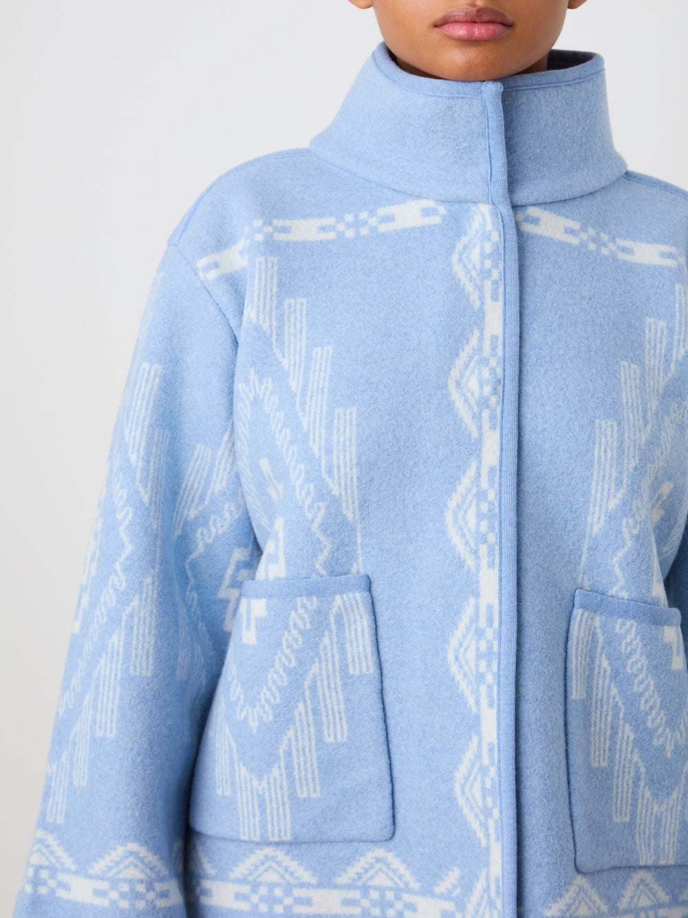A person wears the We Norwegians Pioneer Jacket women - Pre Spring 26, a light blue Merino wool piece with white geometric patterns, high collar, and two front pockets. Image crops at shoulders and mid-thigh.