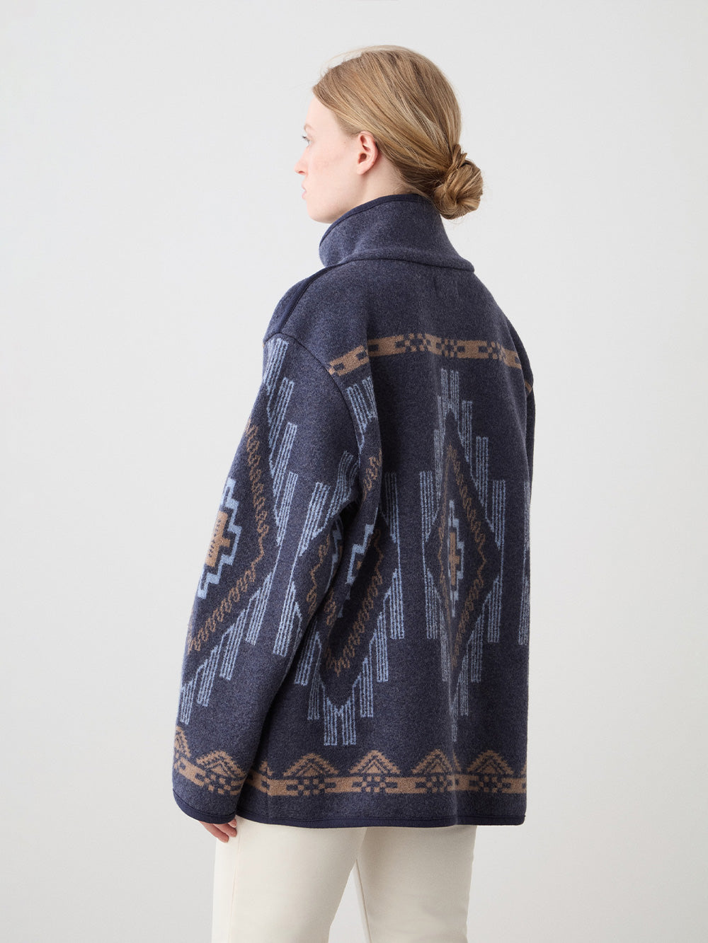 A woman with light brown hair in a low bun wears the We Norwegians Kautokeino Jacket, featuring dark blue and brown boiled wool with a geometric jacquard pattern, standing against a plain light background and facing away from the camera.