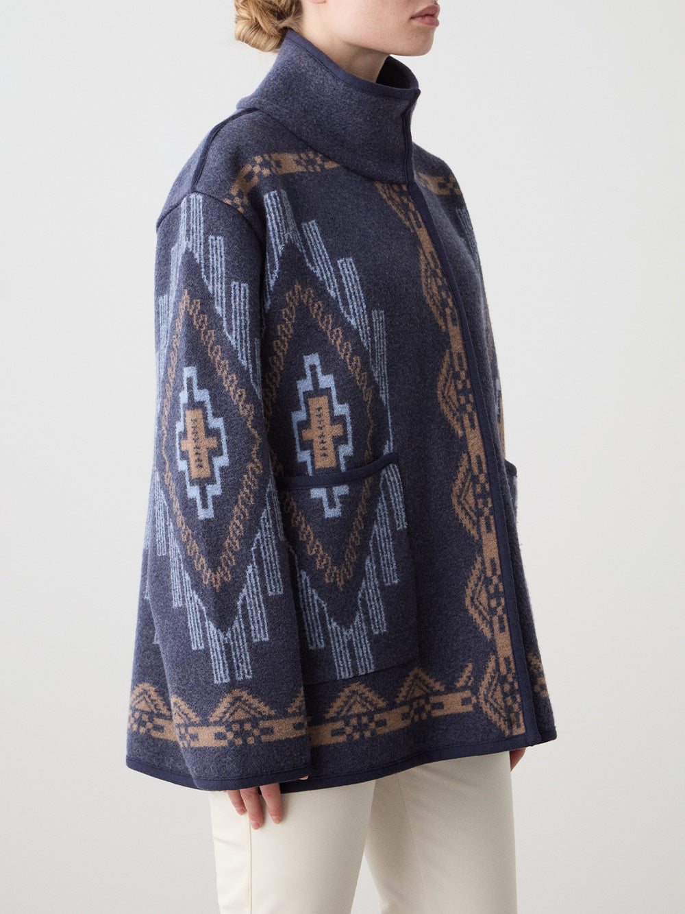 A woman models the We Norwegians Kautokeino Jacket, featuring navy blue boiled wool with geometric brown and light blue jacquard patterns, high collar, and large sleeves. Only the jacket from the shoulders down is visible against a white background.