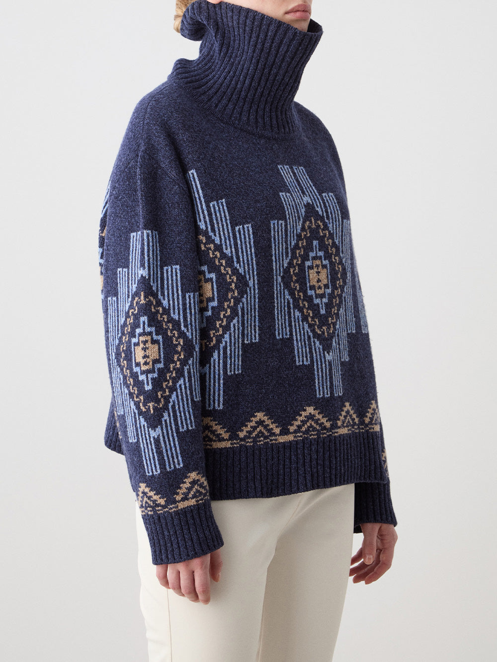 A woman wears the We Norwegians Pioneer Sweater in navy with light blue and beige geometric patterns, paired with off-white pants, standing against a plain, light background.