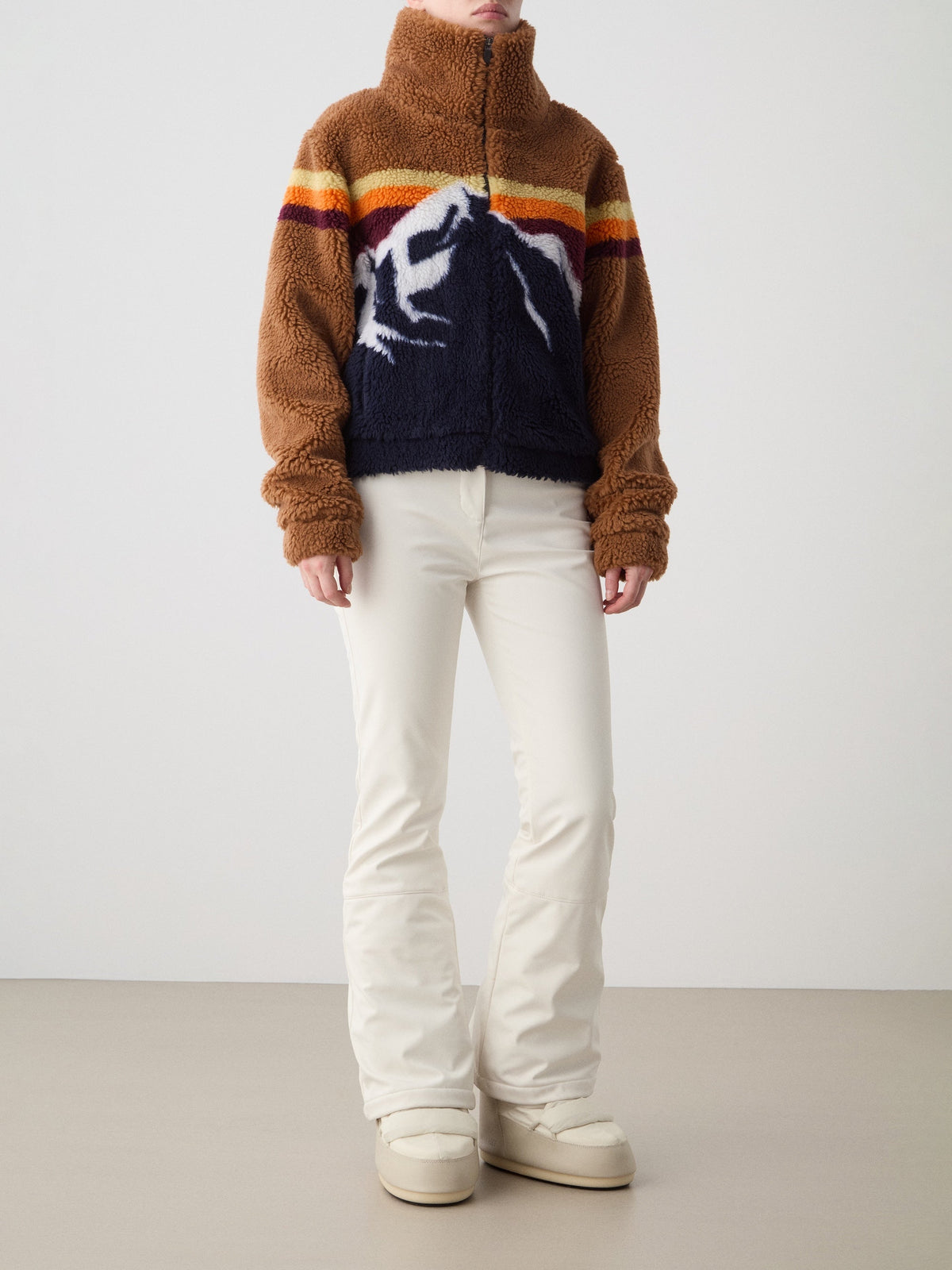 A woman models the We Norwegians Snowmass Jacket—an après-ski essential wool-blend outerwear piece featuring a mountain pattern in fuzzy brown and navy—styled with cream-colored pants and white shoes, set against a plain light background.