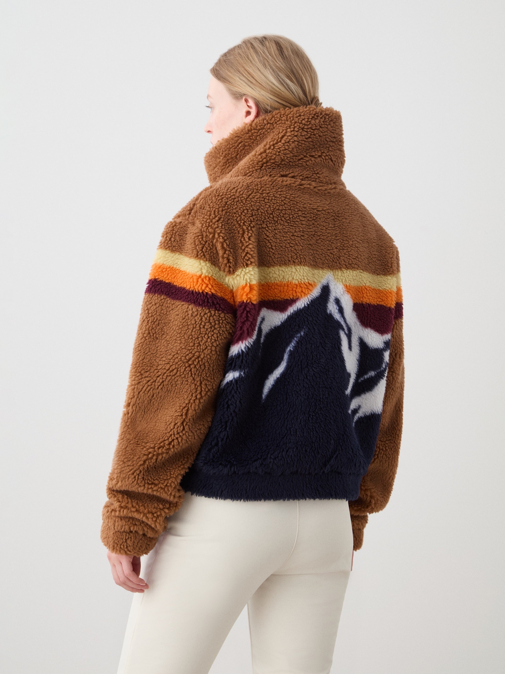 A person with light hair wears the We Norwegians Snowmass Jacket for women—a brown jacquard fleece with a high collar and mountain motif on the back—styled with white pants, standing against a plain light background.