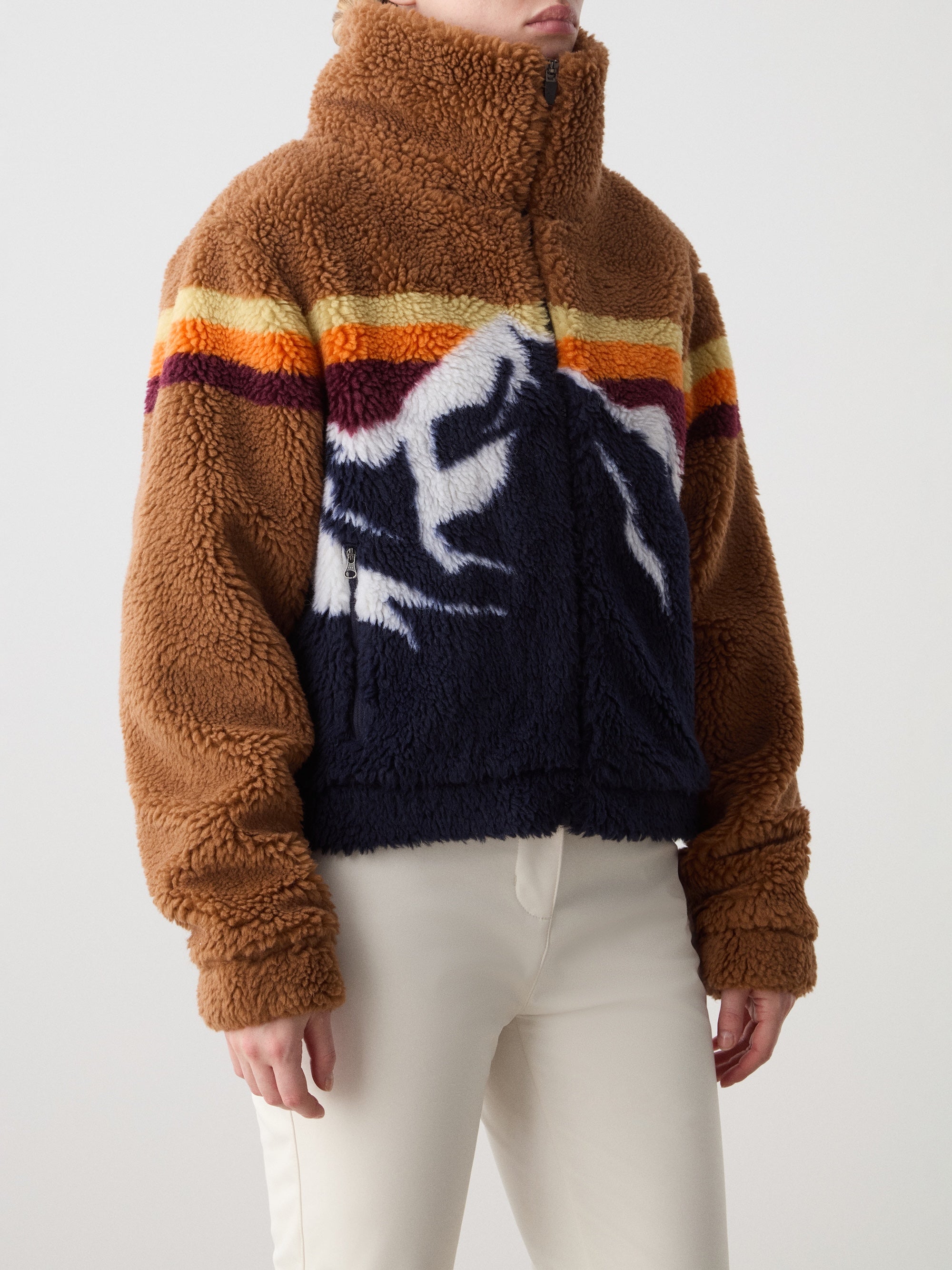 A woman models the We Norwegians Snowmass Jacket—a brown and navy jacquard fleece with colorful stripes and a mountain motif—paired with off-white pants for the perfect après-ski look against a simple background.