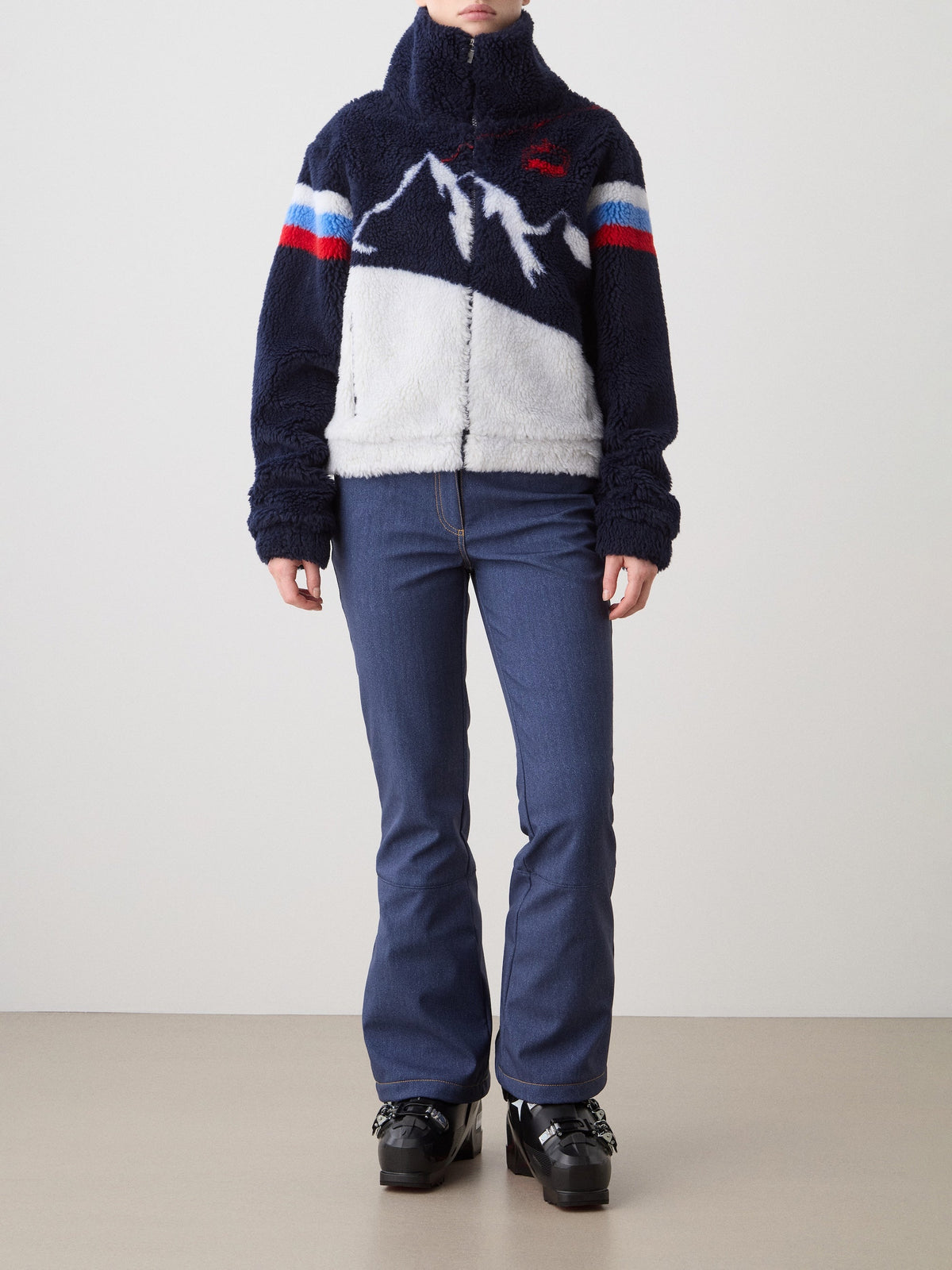 A person stands against a plain light background wearing blue jeans, black platform shoes, and the We Norwegians Snowmass Jacket for women—a fuzzy navy wool-blend jacket with a mountain design, red and blue sleeve stripes, and a high collar—making it the perfect après-ski essential.