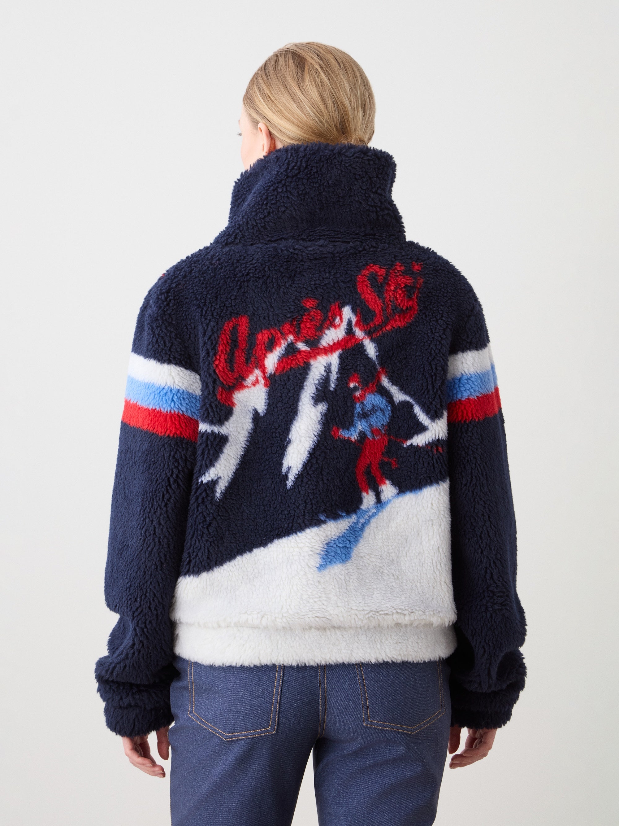 A blonde woman faces away, wearing the We Norwegians Snowmass Jacket: a navy jacquard fleece with red, white, and blue sleeve stripes. The back shows a skiing graphic and "Après Ski" in red—perfect for après-ski style.