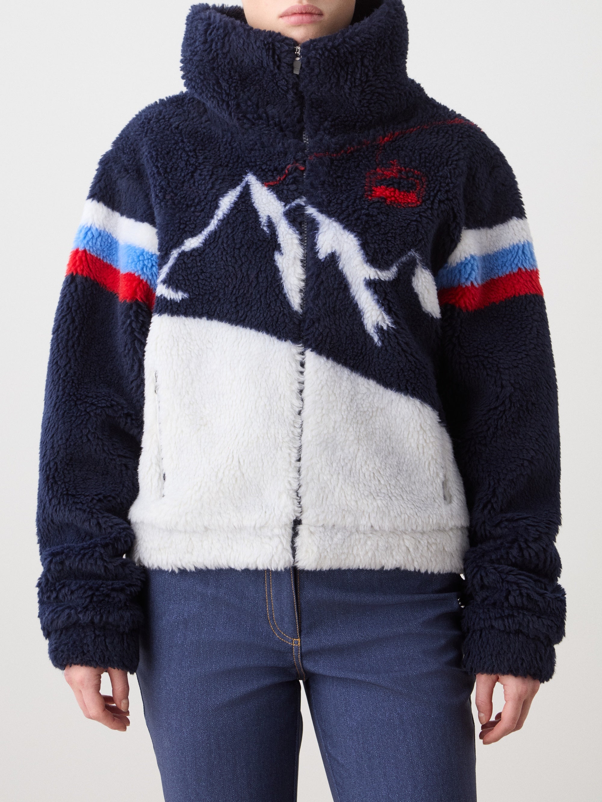 A person wears the We Norwegians Snowmass Jacket: a fuzzy navy and white jacquard fleece zip-up with a high collar, mountain pattern, and red, white, and blue chest and sleeve stripes, paired with blue jeans.