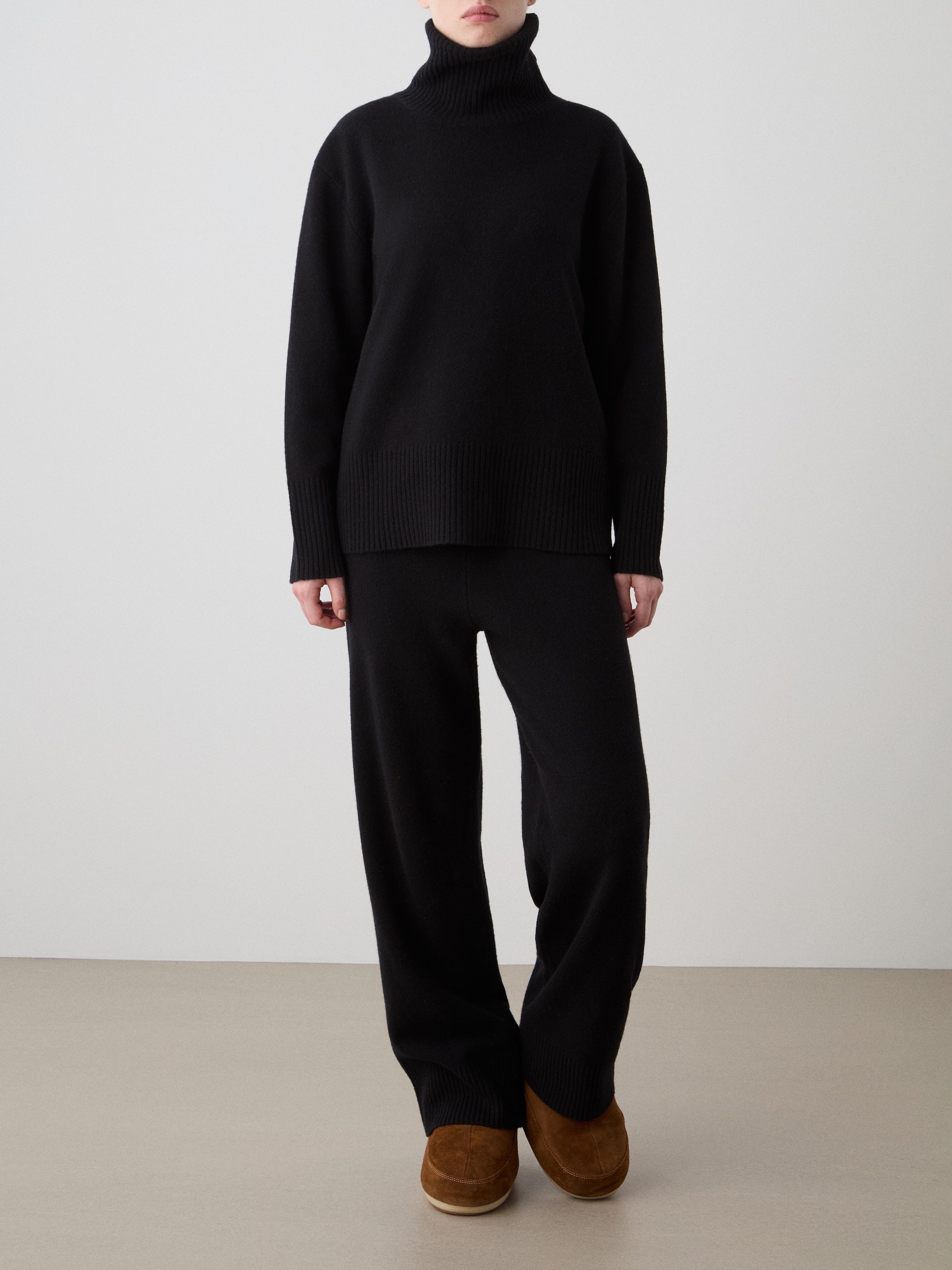 A person stands against a plain background wearing the We Norwegians Geilo Jogger for women, paired with a loose black turtleneck and brown slip-on shoes. Their face is not visible and their hands are relaxed by their sides.