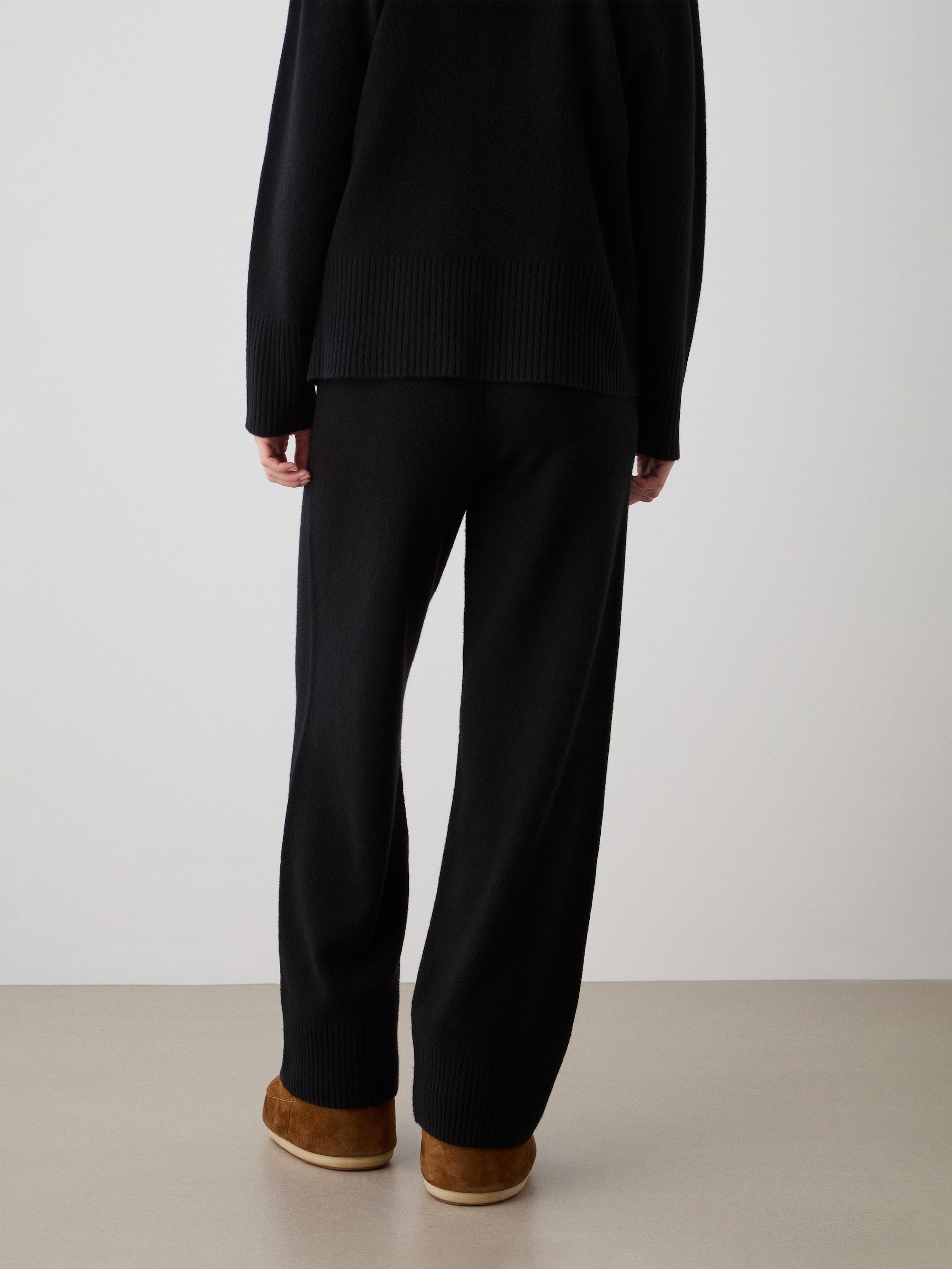 A person stands facing away, wearing We Norwegians' Geilo Jogger women in black, paired with a black long-sleeve top and brown slip-on shoes with cream soles, on a beige floor against a light background.