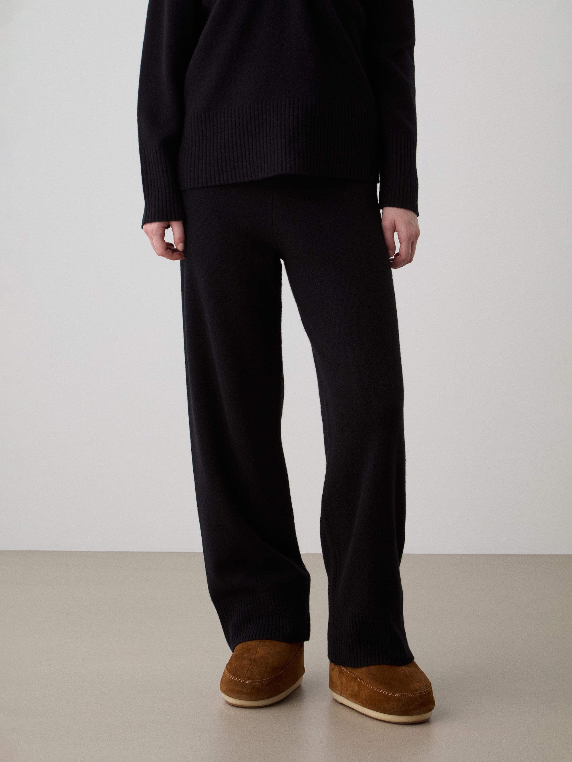 A person stands indoors wearing We Norwegians Geilo Jogger women pants, a black long-sleeve sweater, and brown slip-on shoes. The photo is cropped at the shoulders, so their face isn’t visible.