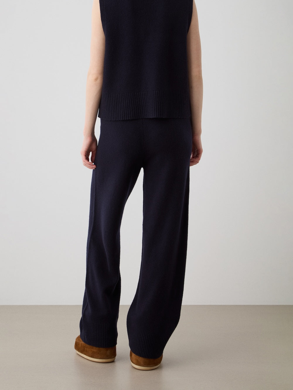 A person stands facing away, wearing We Norwegians' Geilo Jogger women—sleeveless navy knit top with matching relaxed-fit wide-leg pants—and brown slippers, against a plain light-colored wall and floor.