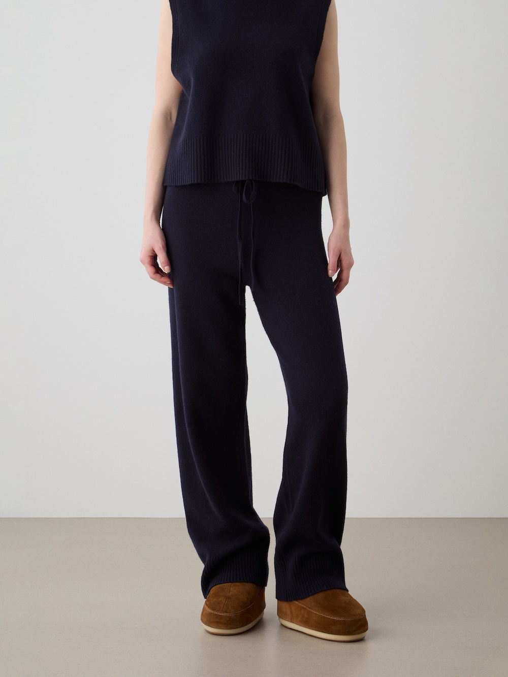 A person (head not visible) stands against a plain background, wearing We Norwegians Geilo Jogger women’s relaxed fit navy knit pants with a sleeveless dark navy knit top and brown suede slippers.