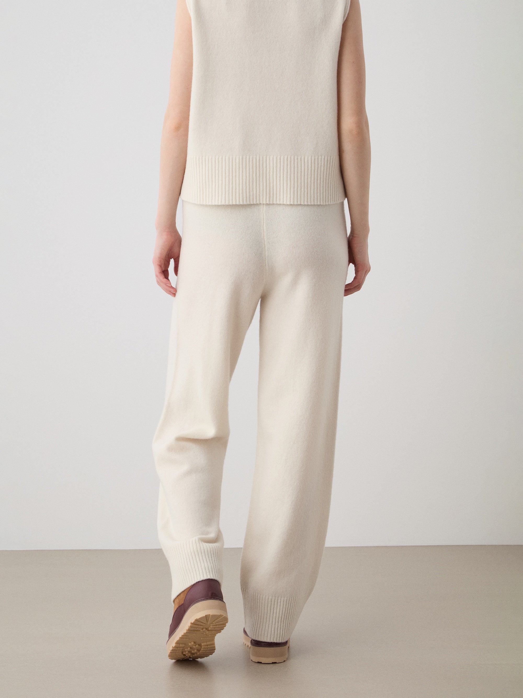A person faces away in a minimal indoor space, wearing We Norwegians' Geilo Jogger women knit pants with an off-white sleeveless top and brown shoes, standing on a light floor against a plain wall.
