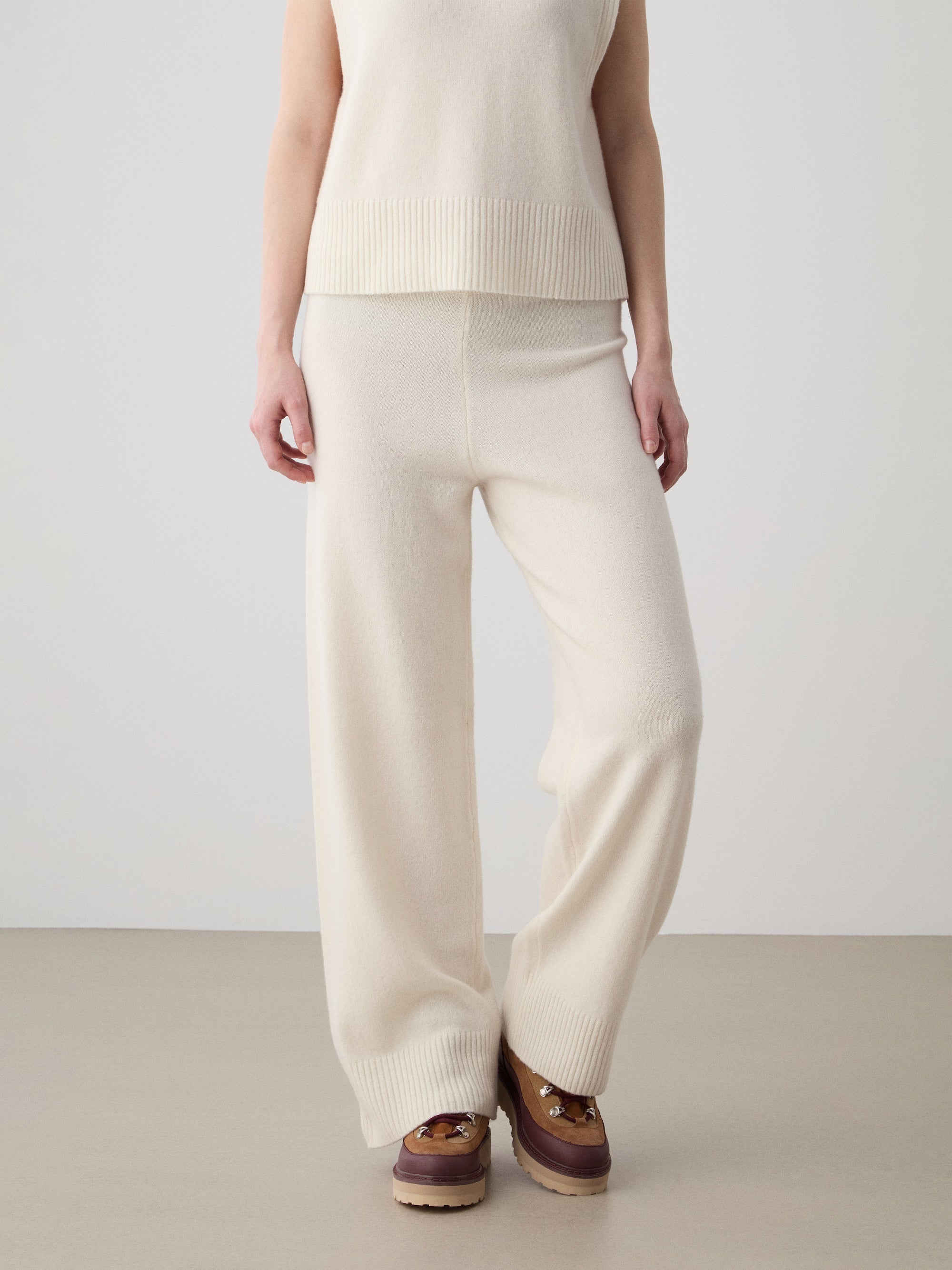 A person models the Geilo Jogger women by We Norwegians: a sleeveless cream knit top with matching cozy pants, paired with brown platform shoes. The cropped photo shows them from shoulders down against a plain background.