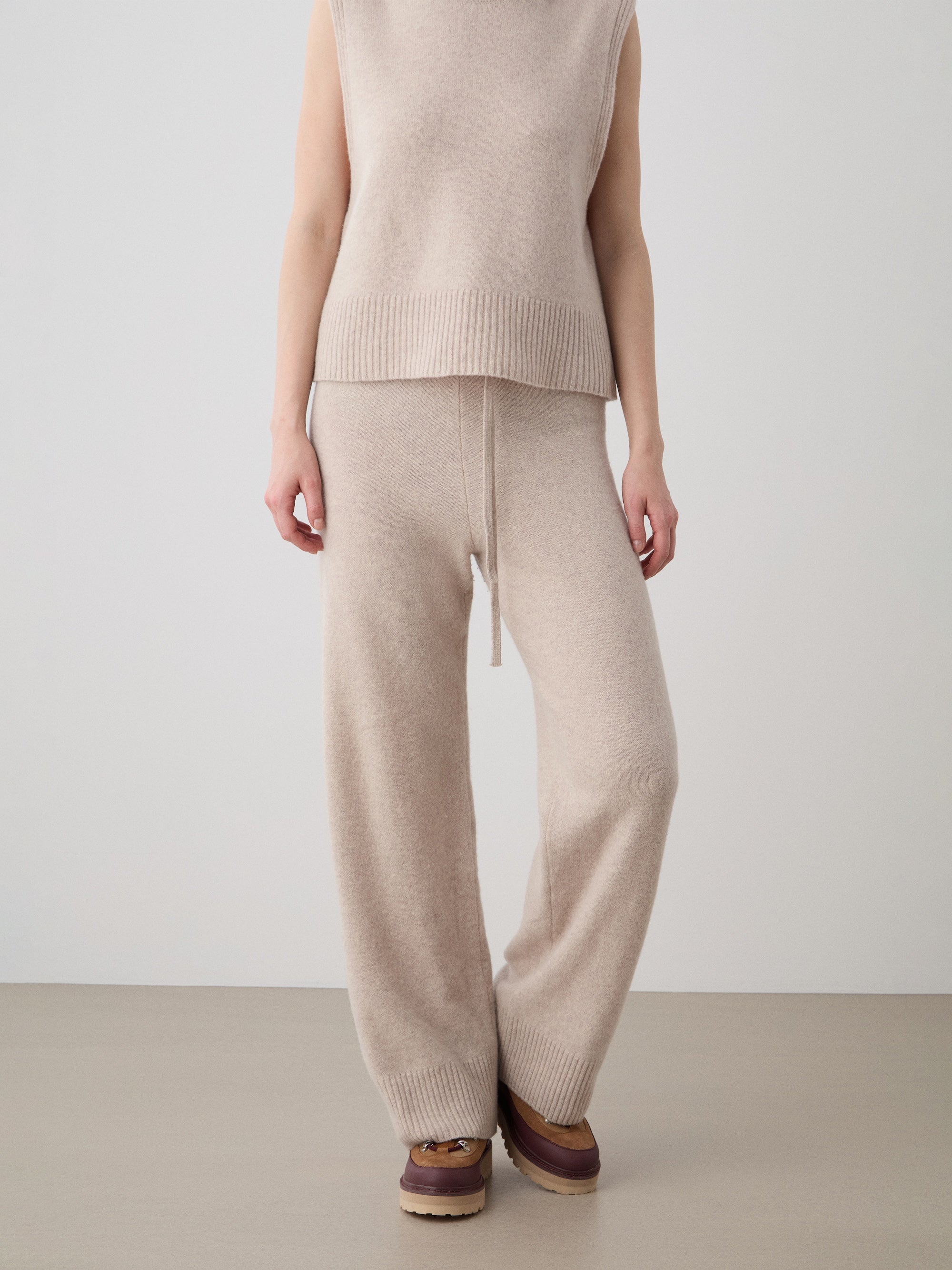 A person stands against a plain light background wearing We Norwegians’ Geilo Jogger women pants, a sleeveless beige knit top, and brown slip-on shoes.