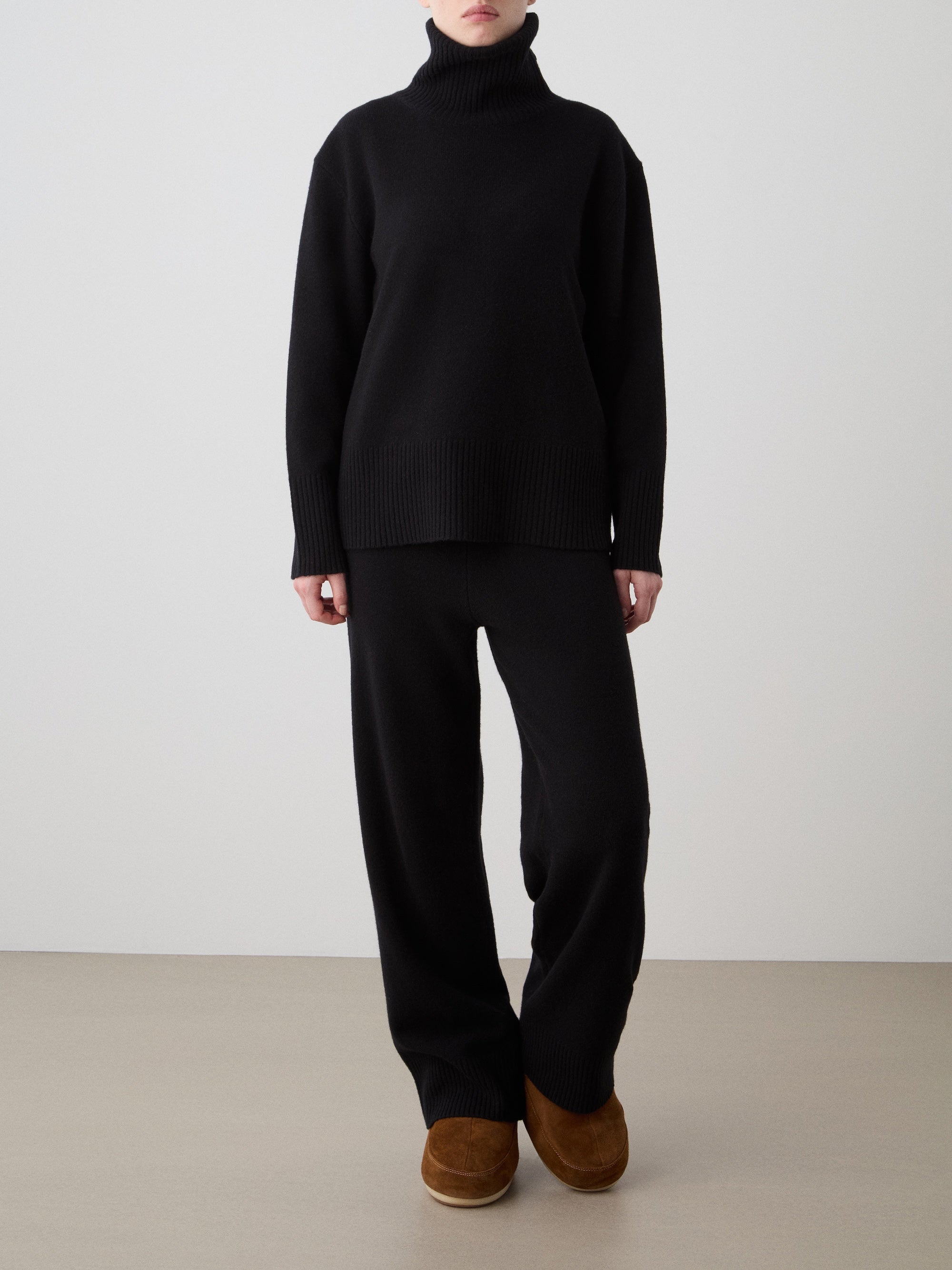 A person stands against a plain background wearing the We Norwegians Geilo BoyFriend Sweater for women in black, paired with matching black pants and brown slippers. Their hands rest at their sides, and their face is not visible.