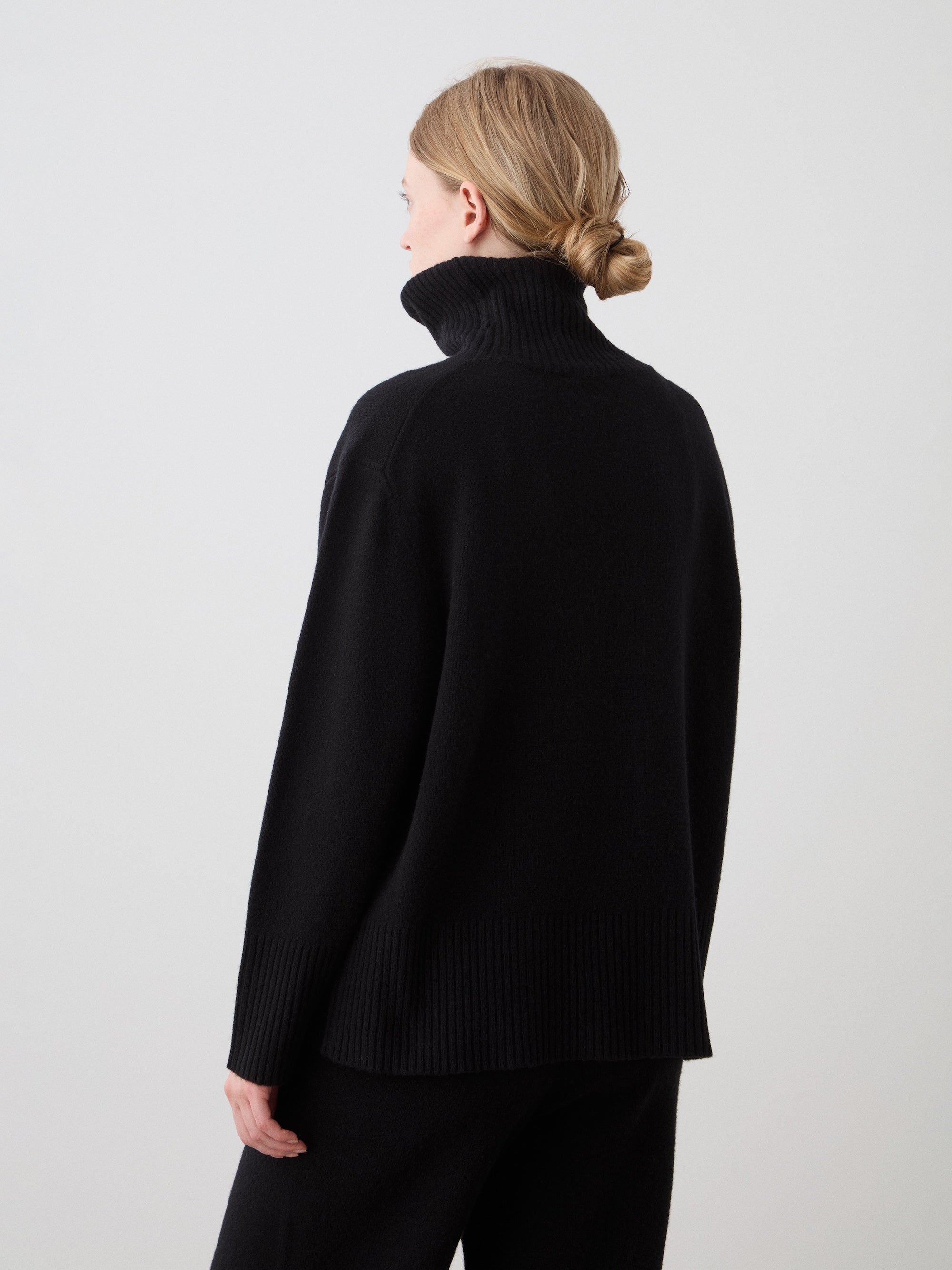 A woman with light hair in a low bun is seen from behind, wearing the We Norwegians Geilo BoyFriend Sweater for women in Extrafine Merino Wool with black pants, standing against a plain light background.