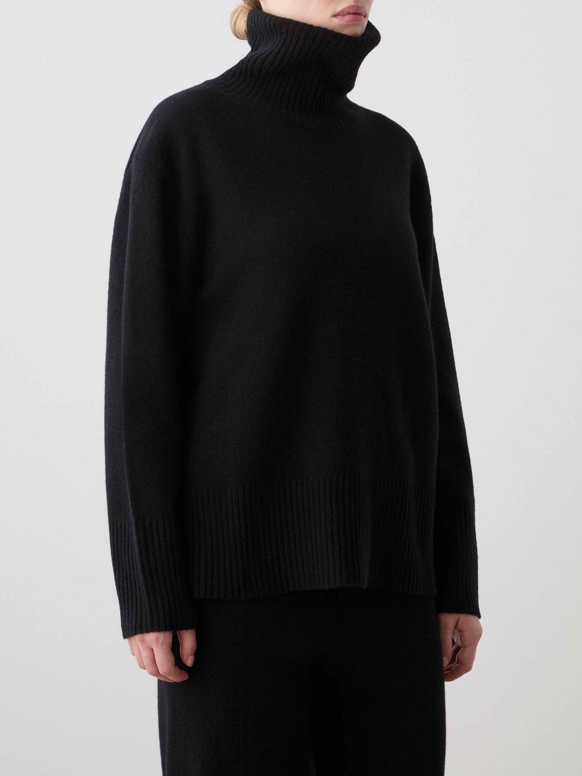 A person in the We Norwegians Geilo BoyFriend Sweater for women and matching black pants stands against a plain white background. The loose black turtleneck has wide ribbed cuffs and hem, with the wearer's face partially out of frame.